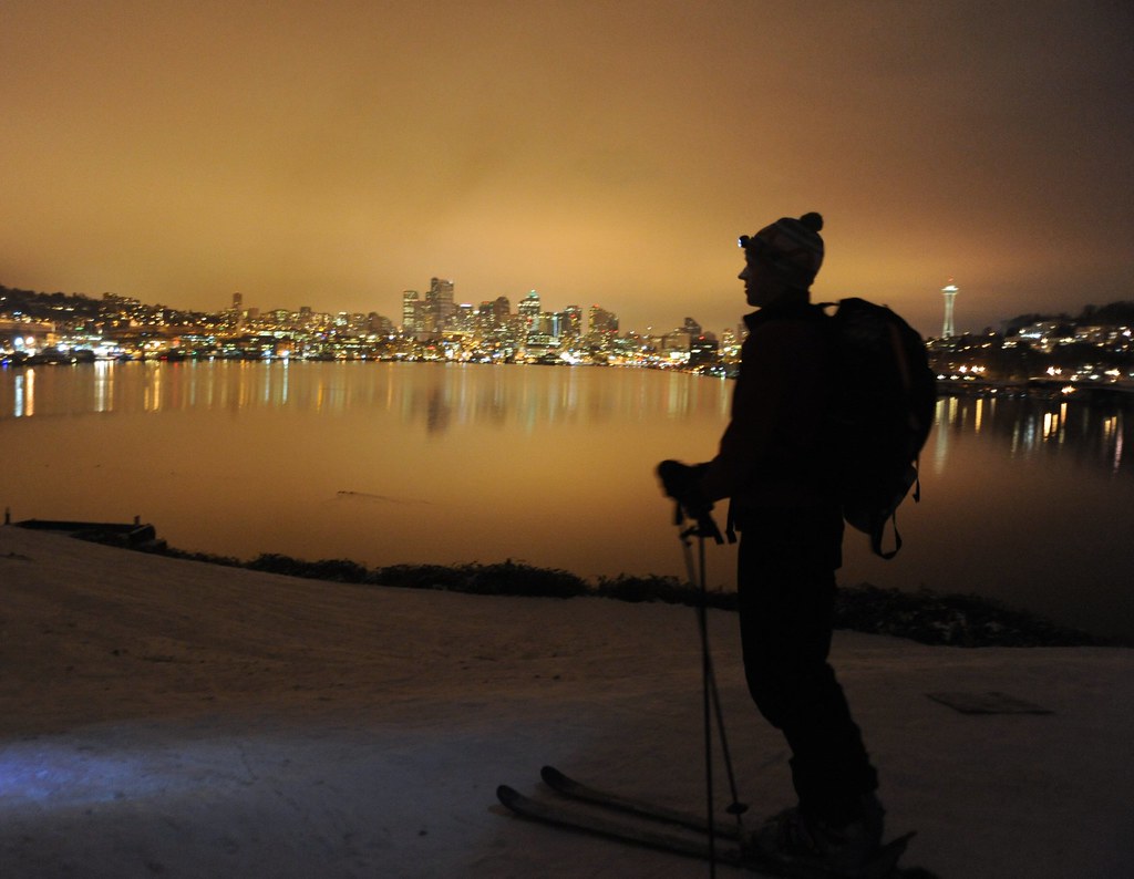 On record snow, night skier with pack and headlight Klarus, city reflected in Lake Union, Space Needle, from the top of the hill, USA