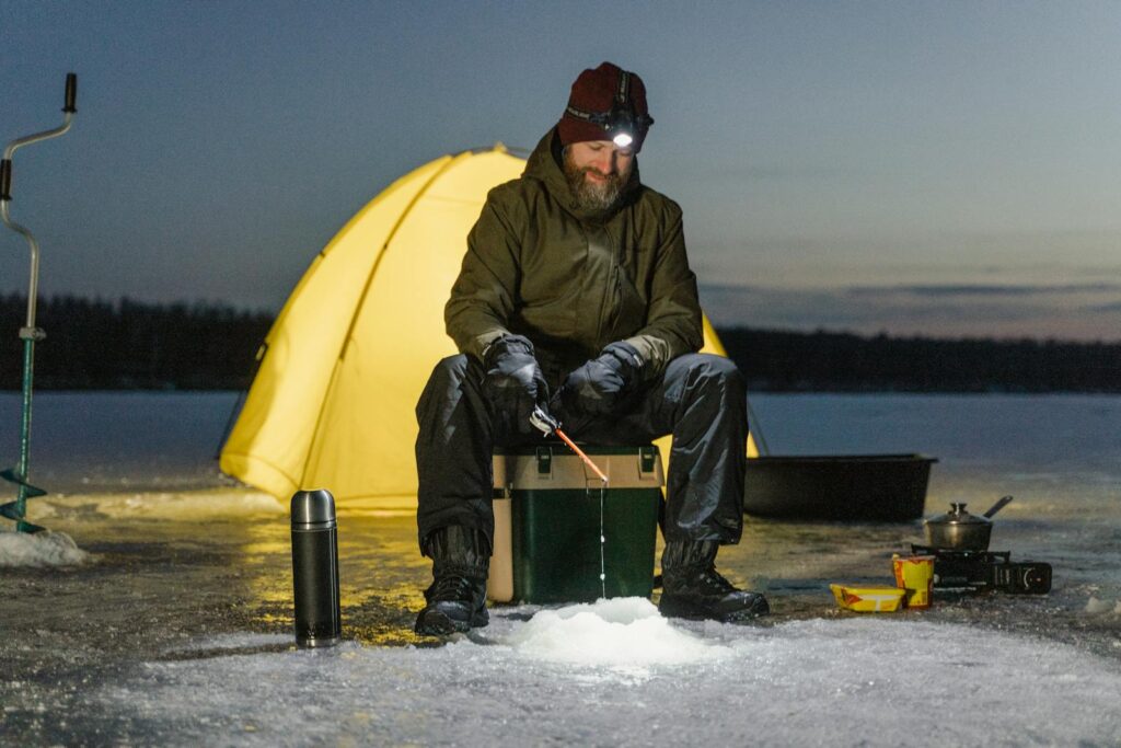 A man ice fishing by tent on a frozen lake during winter night, capturing outdoor adventure with head torch.